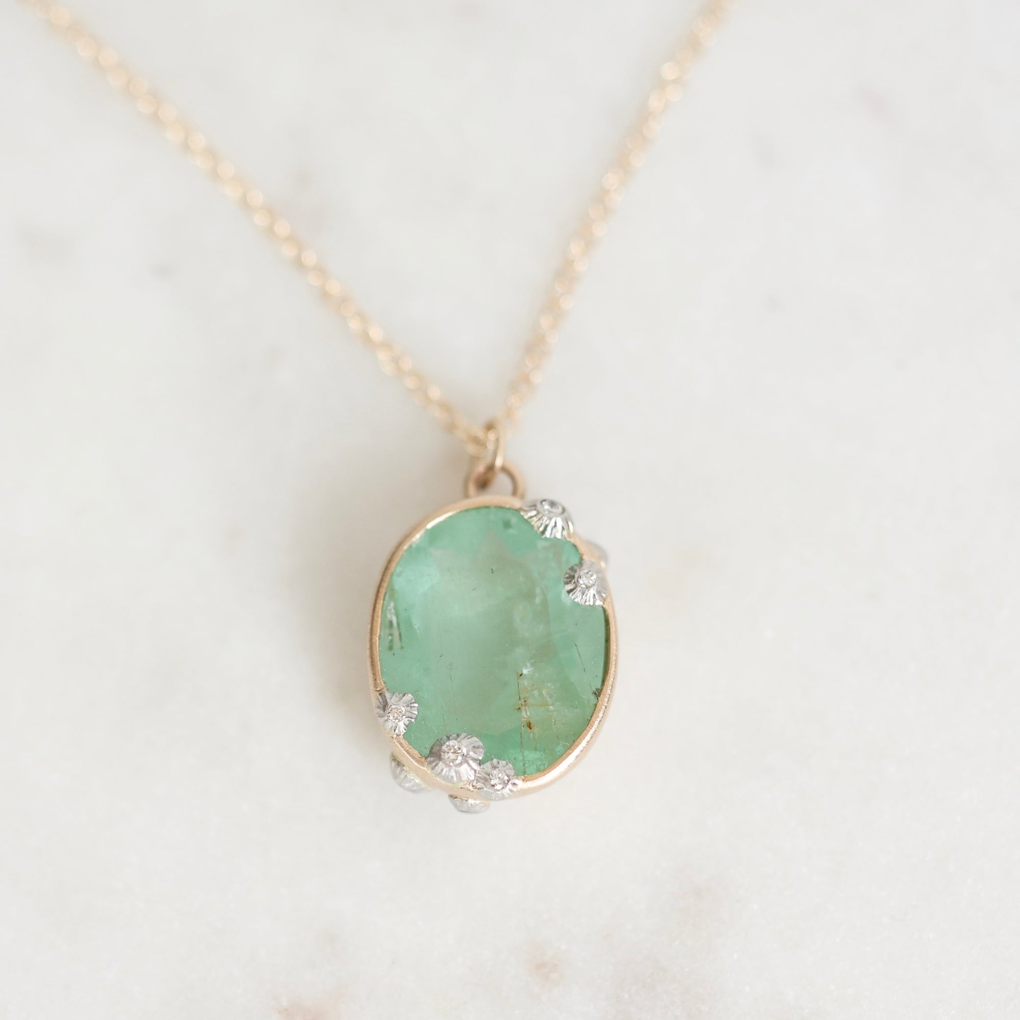 An oval pendant necklace featuring a light green stone in a gold bezel setting with small silver accents, resting on a white marble surface.