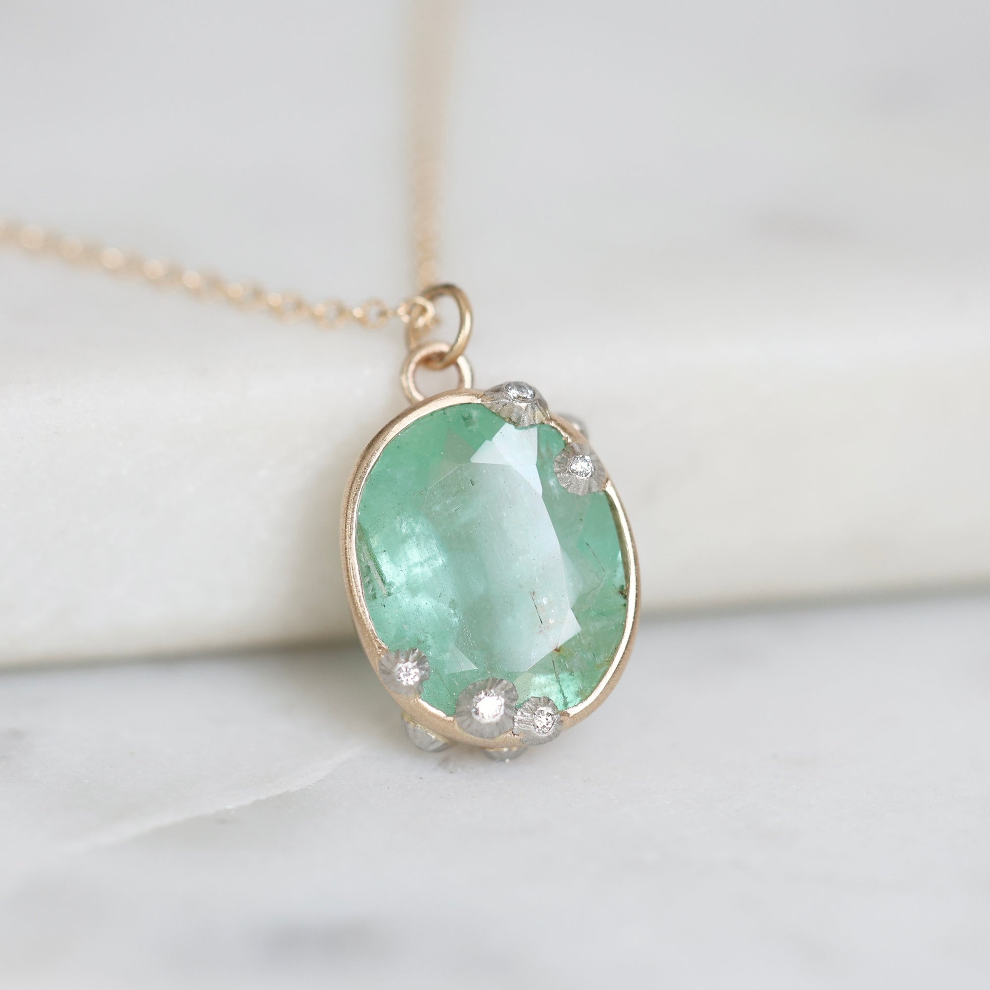 An oval pendant necklace featuring a light green gemstone in a gold bezel setting, accented with small clear stones, resting on a white marble surface.