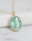 An oval pendant necklace featuring a light green gemstone in a gold bezel setting, accented with small clear stones, resting on a white marble surface.