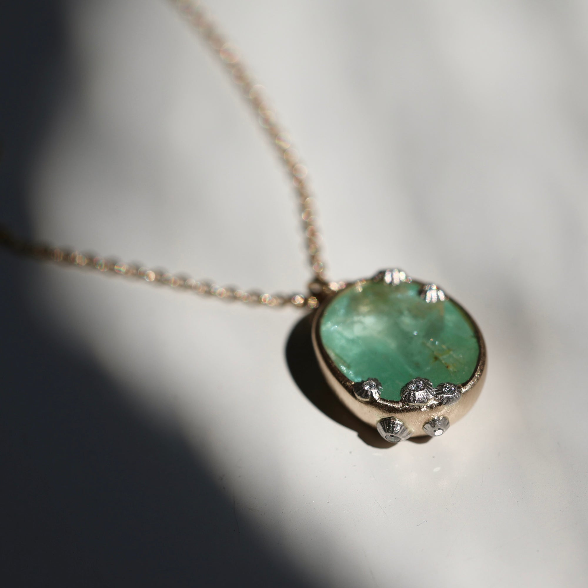 A close-up of a gold chain necklace with an oval pendant featuring a green stone and small clear accent stones, photographed on a white surface with strong sunlight and shadows.