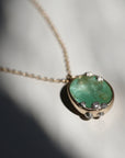 A close-up of a gold chain necklace with an oval pendant featuring a green stone and small clear accent stones, photographed on a white surface with strong sunlight and shadows.