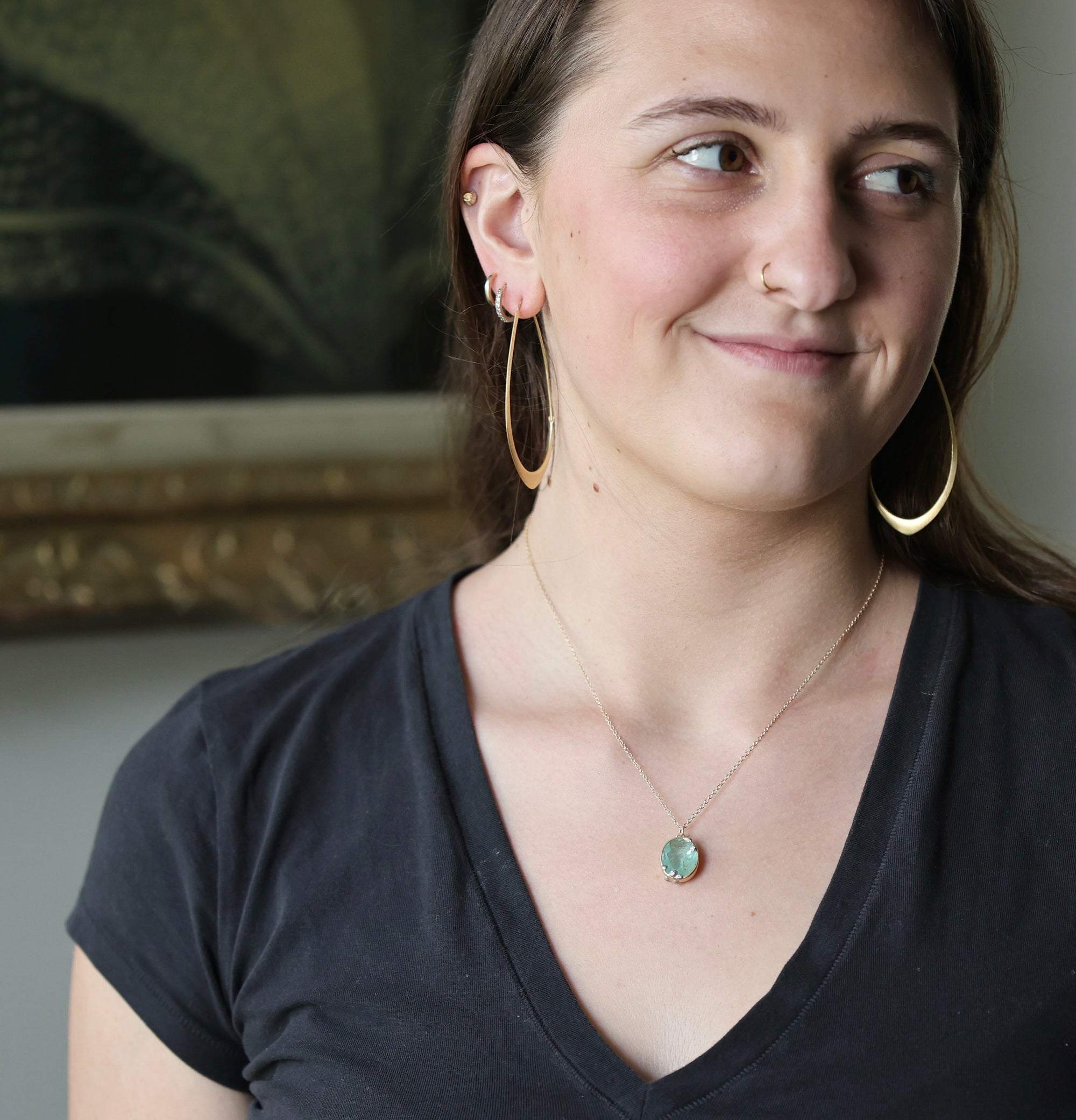A woman wearing a delicate gold chain necklace with a round, light green stone pendant, styled with large gold hoop earrings and a black V-neck t-shirt.
