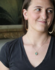 A woman wearing a delicate gold chain necklace with a round, light green stone pendant, styled with large gold hoop earrings and a black V-neck t-shirt.