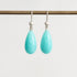 A pair of silver drop earrings with teardrop-shaped light turquoise blue stones hanging from a horizontal display rod against a plain light gray background.