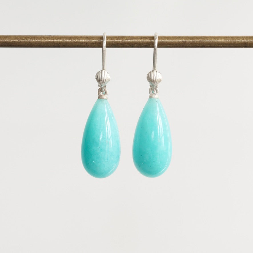 A pair of silver drop earrings with teardrop-shaped light turquoise blue stones hanging from a horizontal display rod against a plain light gray background.