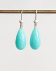 A pair of silver drop earrings with teardrop-shaped light turquoise blue stones hanging from a horizontal display rod against a plain light gray background.
