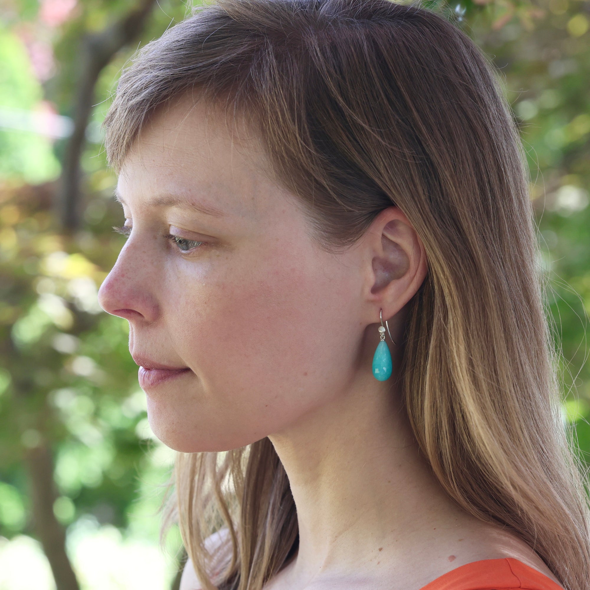 A woman in profile wearing a small turquoise teardrop dangle earring with a silver hook, set against a soft-focus outdoor background of green foliage.