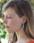 A woman in profile wearing a small turquoise teardrop dangle earring with a silver hook, set against a soft-focus outdoor background of green foliage.