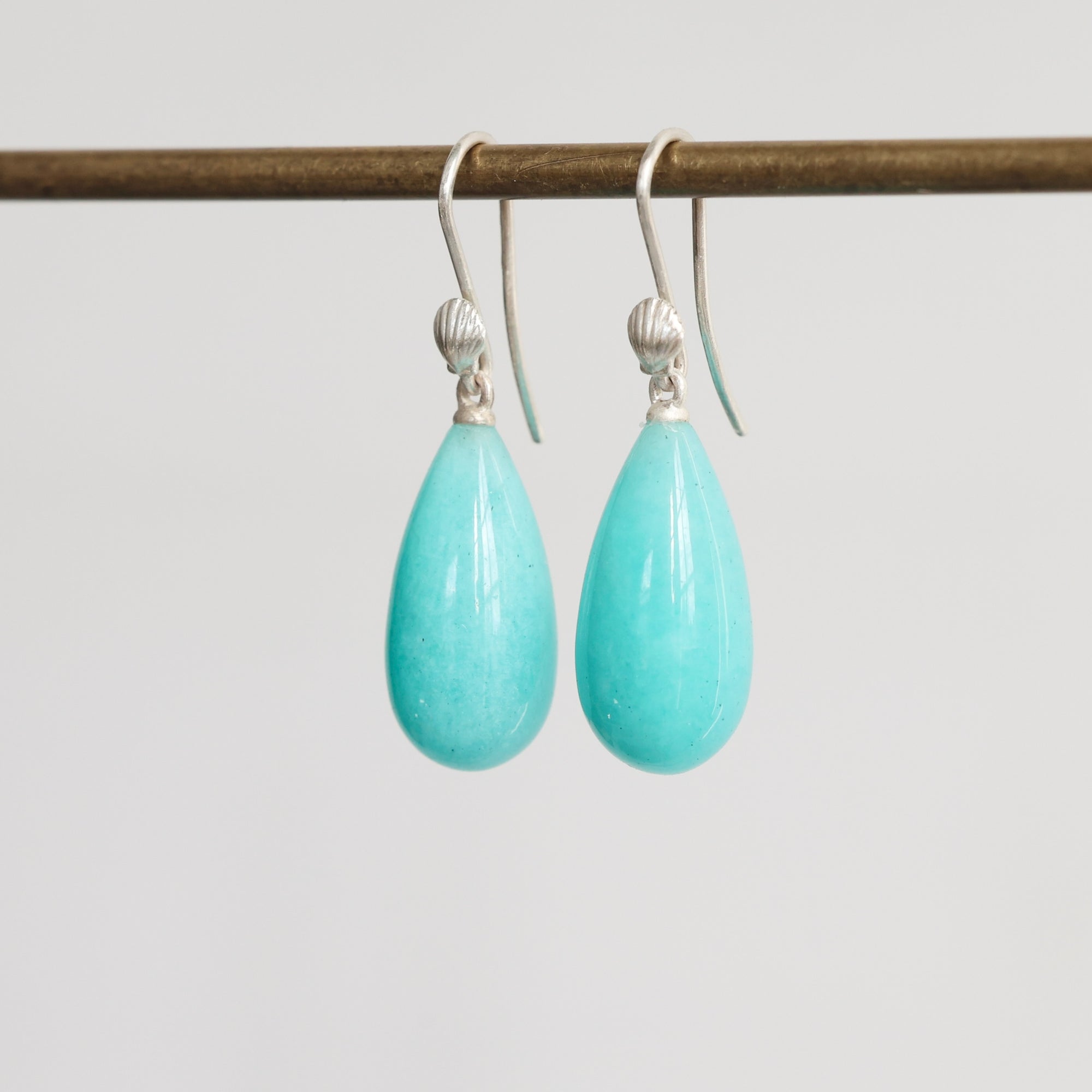 A pair of teardrop-shaped turquoise drop earrings with silver hooks, hanging from a dark horizontal rod against a light gray background.
