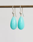 A pair of teardrop-shaped turquoise drop earrings with silver hooks, hanging from a dark horizontal rod against a light gray background.