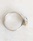 A silver ring with a textured gold setting holding a small oval green gemstone, photographed from above on a light off-white background.