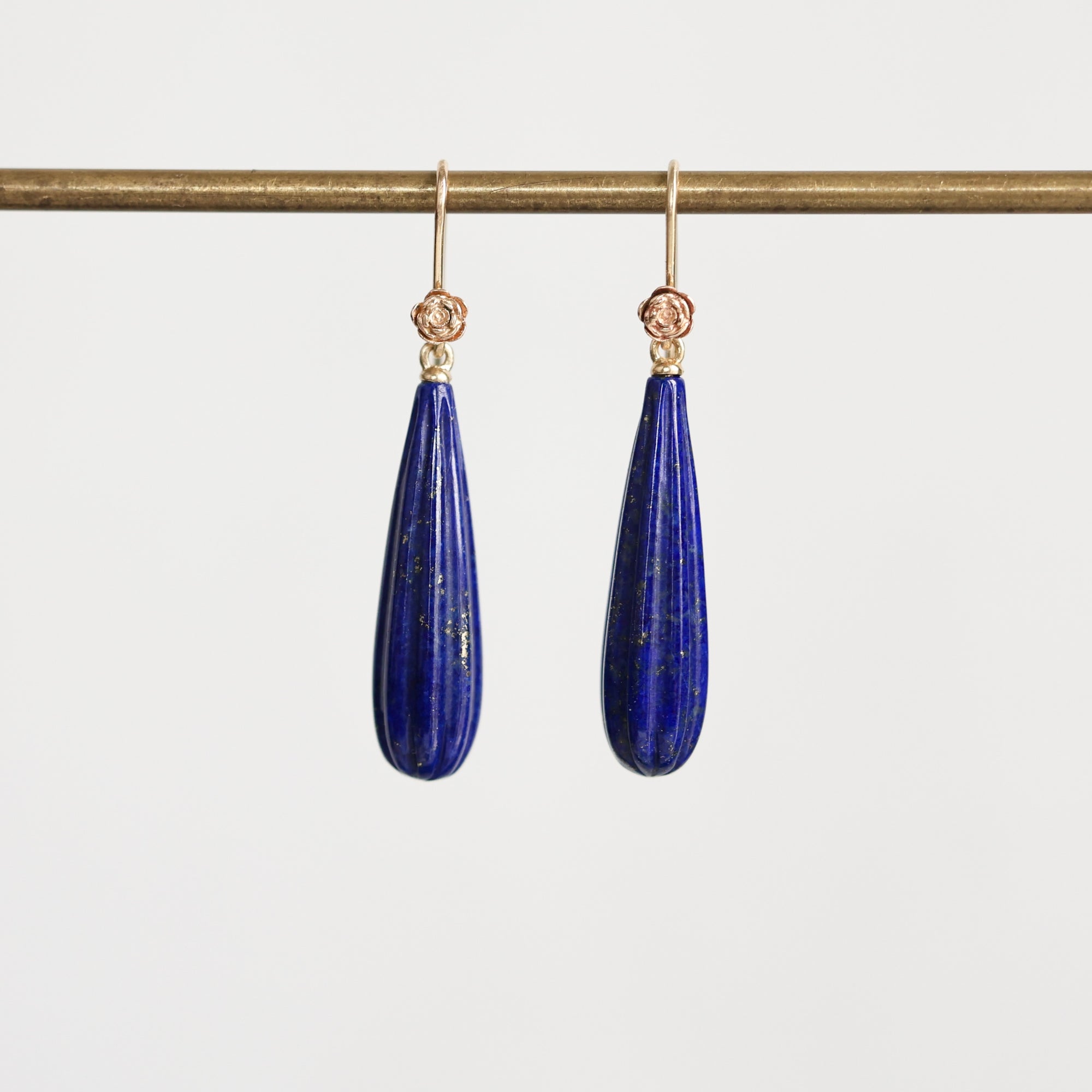 A pair of gold drop earrings with teardrop-shaped dark blue lapis lazuli stones, hanging from a gold rod against a light gray background.