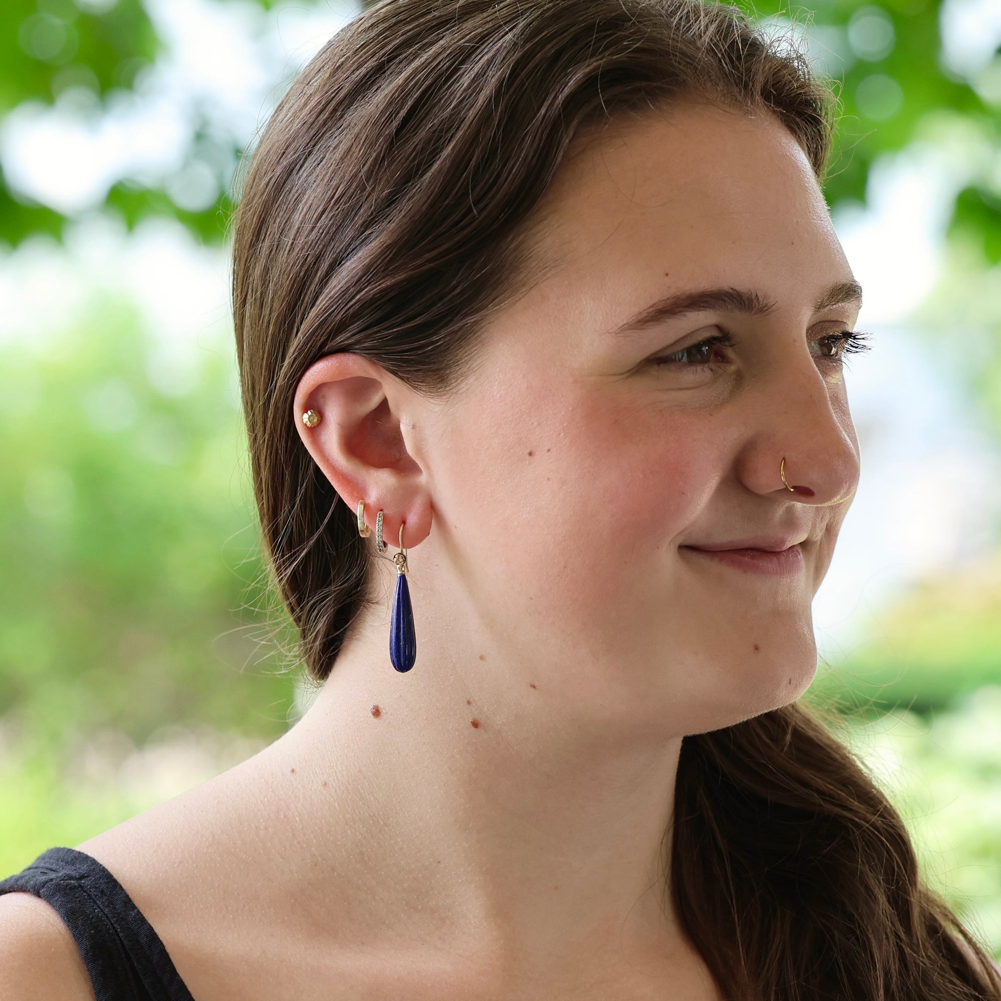 A woman wearing a dark blue teardrop-shaped drop earring with gold hoop and stud earrings, shown in a close-up profile against a blurred green outdoor background.