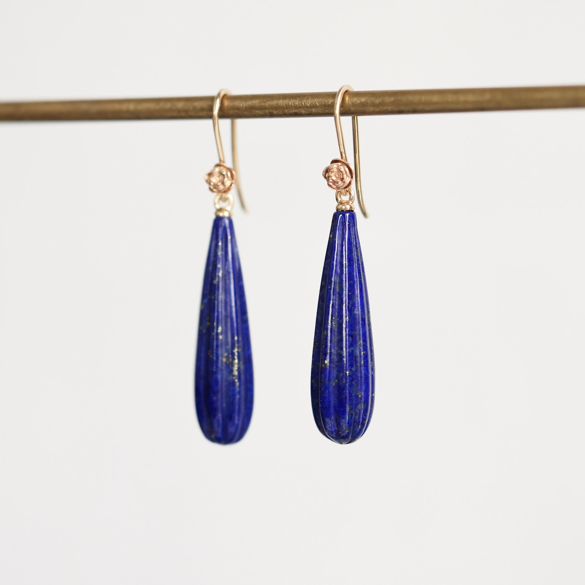 A pair of teardrop-shaped lapis lazuli earrings with gold hooks, hanging from a gold display rod against a light gray background.