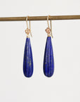 A pair of teardrop-shaped lapis lazuli earrings with gold hooks, hanging from a gold display rod against a light gray background.