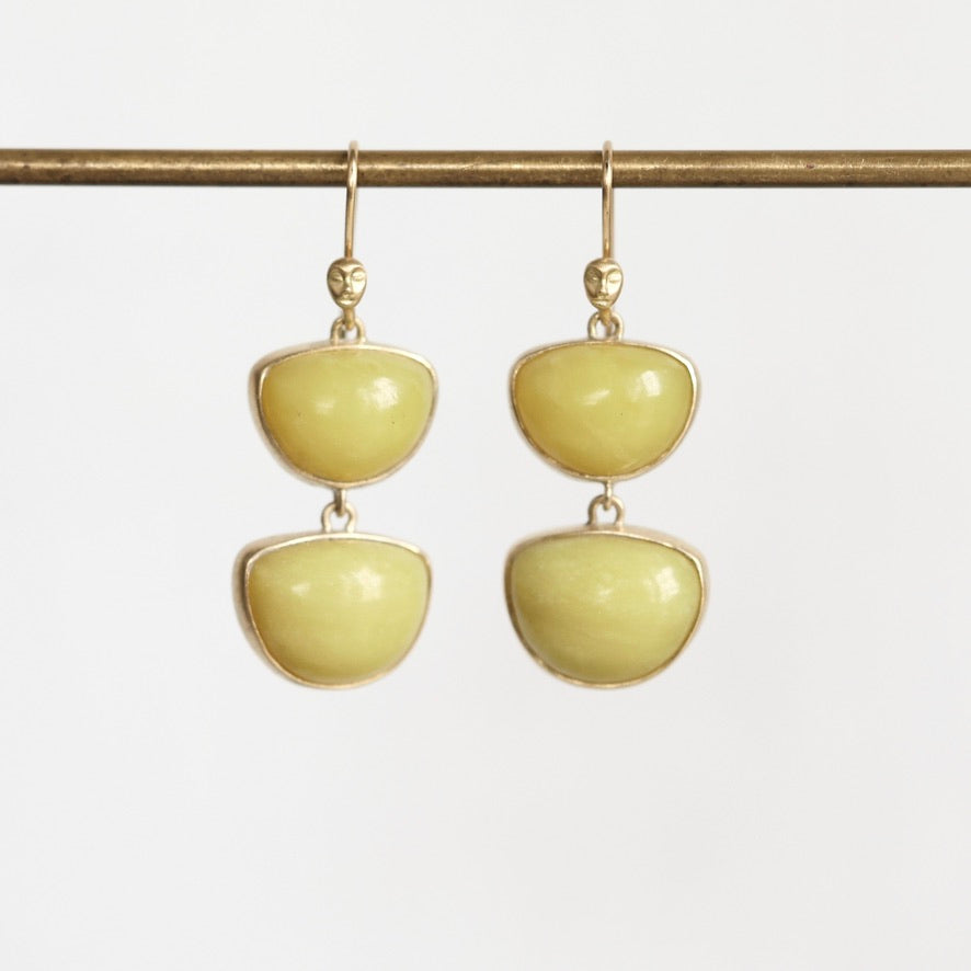 A pair of gold drop earrings with two smooth, rounded pale yellow-green stone segments hanging from gold hooks, displayed against a light gray background.