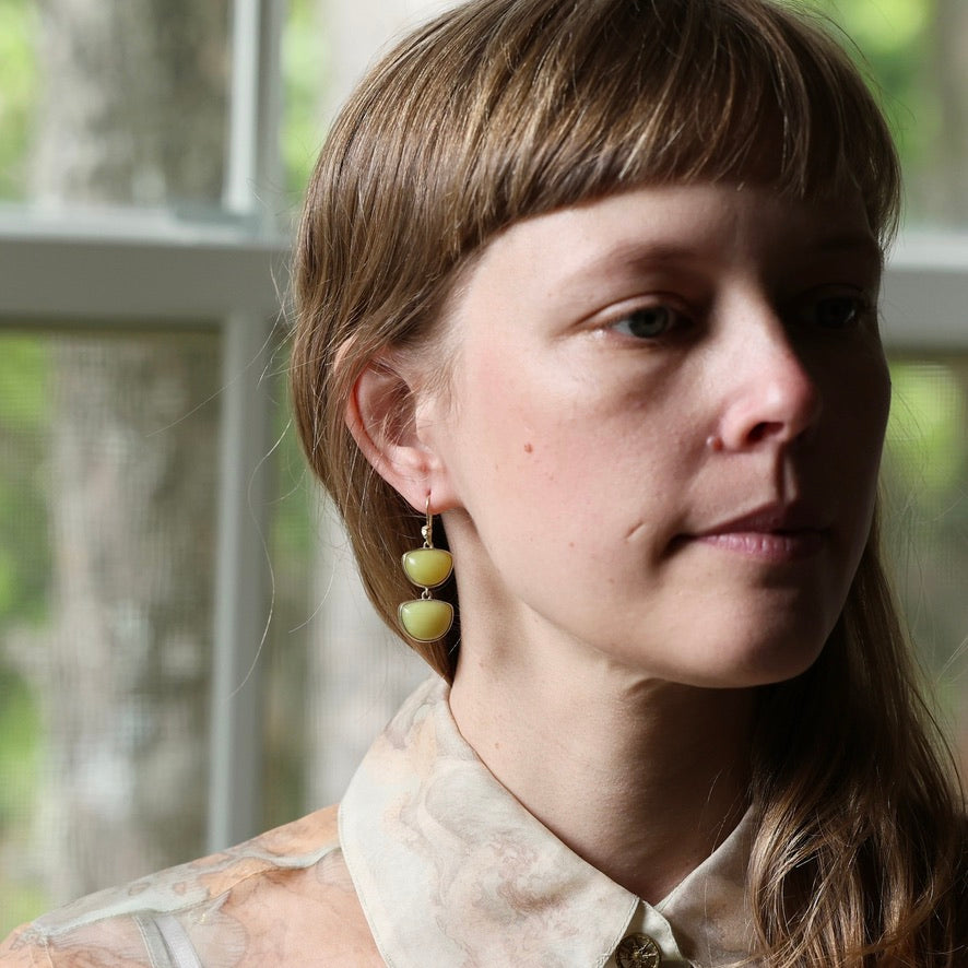 A close-up portrait of a woman wearing a pair of dangle earrings made of two stacked, rounded olive-green beads on a gold-toned hook, set against a soft-focus background of a window.
