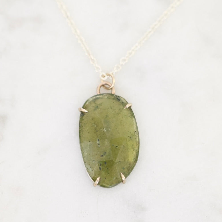 An oval green stone pendant necklace with a gold-toned bezel setting and delicate chain, displayed on a light marble surface.