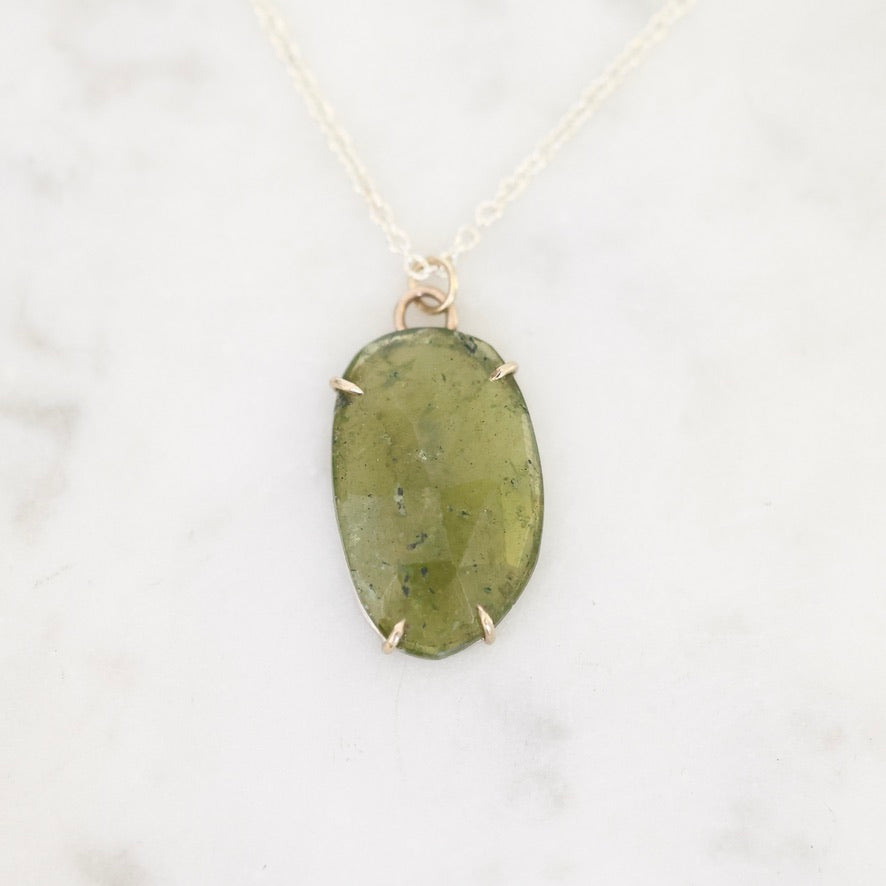 An oval green stone pendant necklace with a gold-toned bezel setting and delicate chain, displayed on a light marble surface.