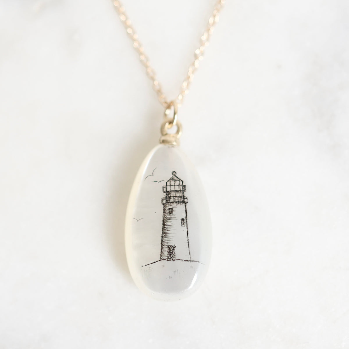 A gold chain necklace with an oval clear resin pendant featuring a black ink sketch of a lighthouse, displayed on a light marble surface.