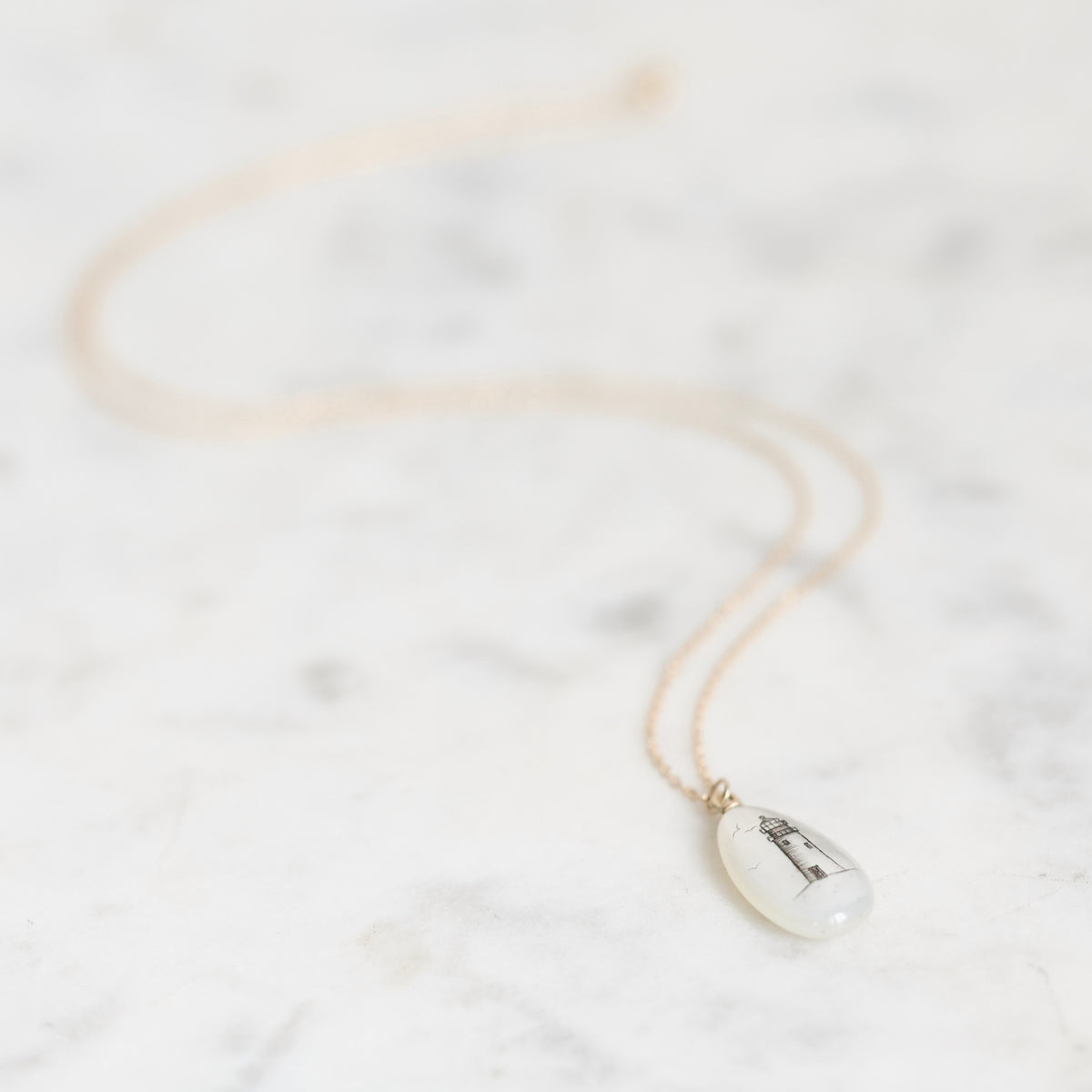 A delicate gold chain necklace with a small, teardrop-shaped white pendant featuring a black lighthouse engraving, displayed on a light marble surface.