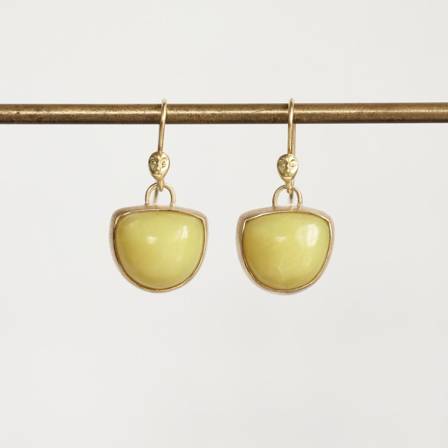 A pair of gold drop earrings with smooth, dome-shaped pale yellow-green stones, hanging from a gold horizontal bar against a light gray background.