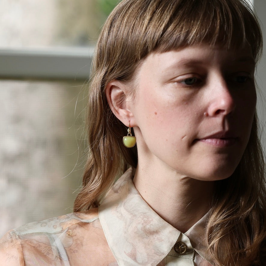 A close-up portrait of a woman wearing a small, round, light green bead earring with a gold-toned top hook, paired with a cream-colored patterned blouse.
