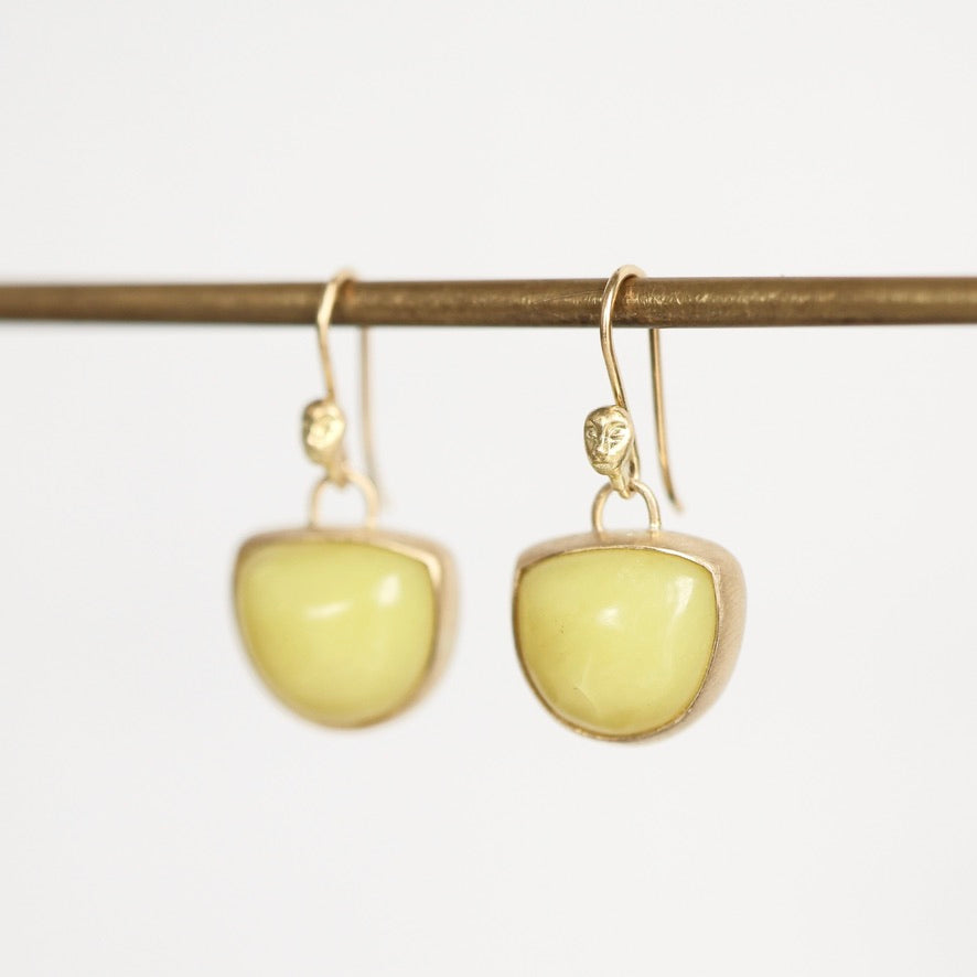 A pair of gold drop earrings with smooth, rounded pale yellow-green stone pendants, hanging from a thin horizontal gold rod against a plain white background.