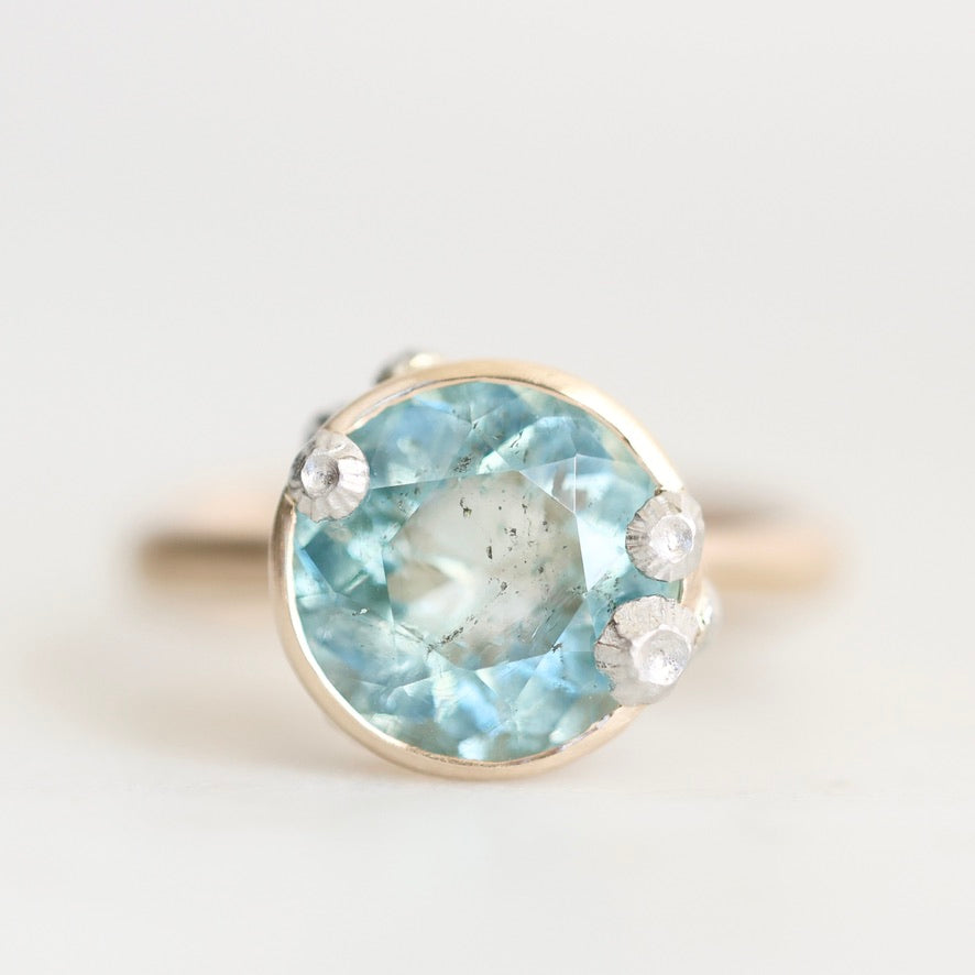 A round-cut light blue gemstone ring in a gold-toned setting, accented with three small clear stones, photographed on a soft light gray background.