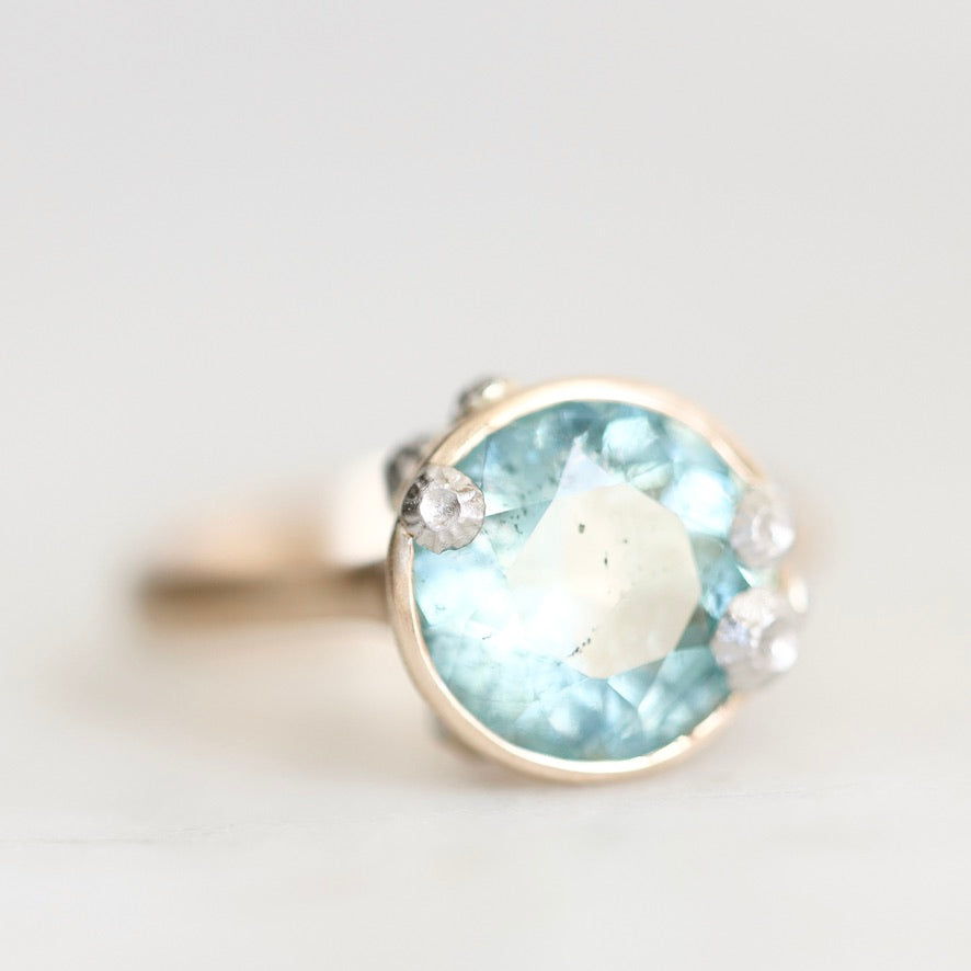 A silver ring featuring a large oval light blue gemstone in a bezel setting, accented with three small round clear stones on the shoulders, photographed against a soft light gray background.