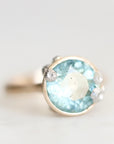 A silver ring featuring a large oval light blue gemstone in a bezel setting, accented with three small round clear stones on the shoulders, photographed against a soft light gray background.