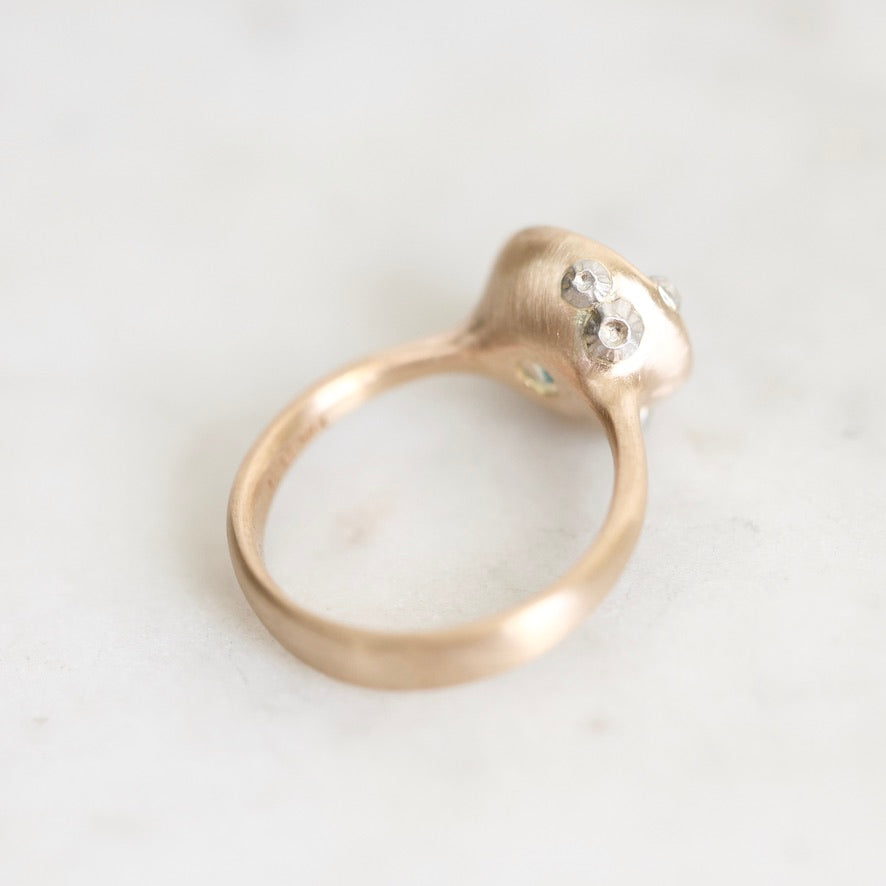 A close-up shot of a gold ring with a textured, organic-shaped setting featuring small clear gemstones, resting on a light gray marble surface.