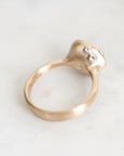 A close-up shot of a gold ring with a textured, organic-shaped setting featuring small clear gemstones, resting on a light gray marble surface.