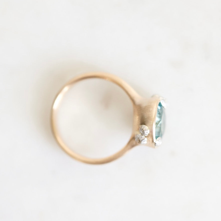 A gold ring with a bezel-set oval light blue gemstone and a small clear accent stone, photographed on a light off-white background.