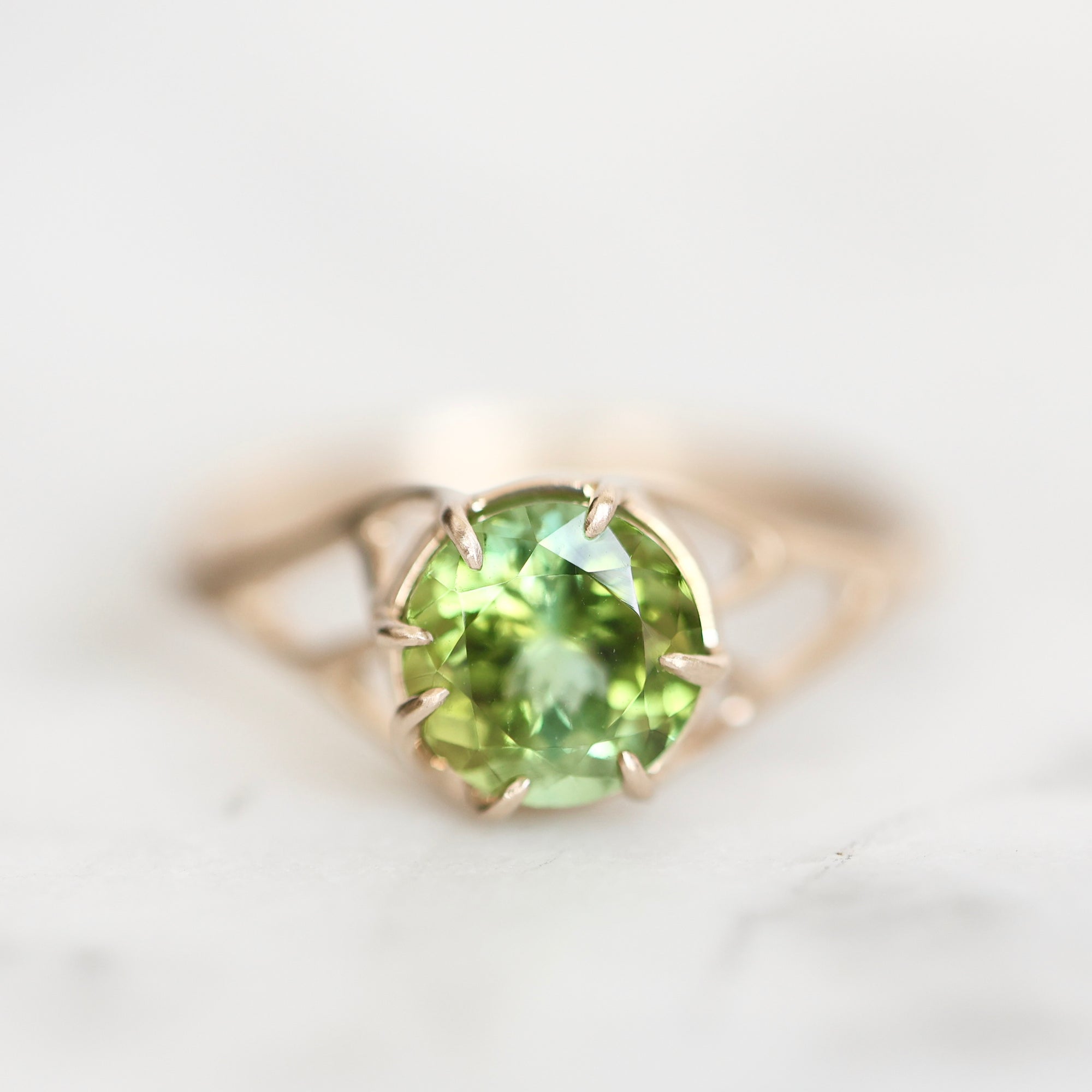 A rose gold ring featuring a round-cut green peridot gemstone in a prong setting, resting on a light marble surface.