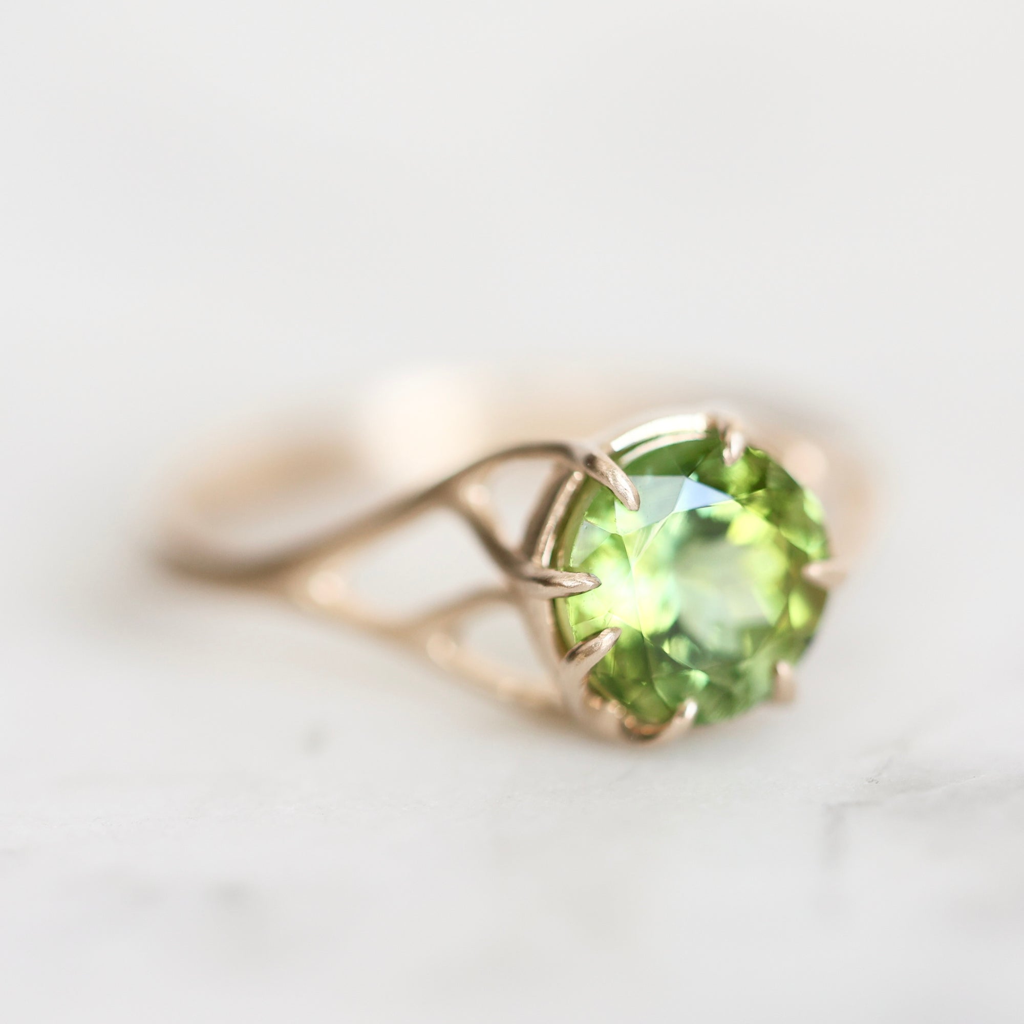 A gold ring featuring a round-cut green peridot gemstone in a prong setting, resting on a light marble surface.