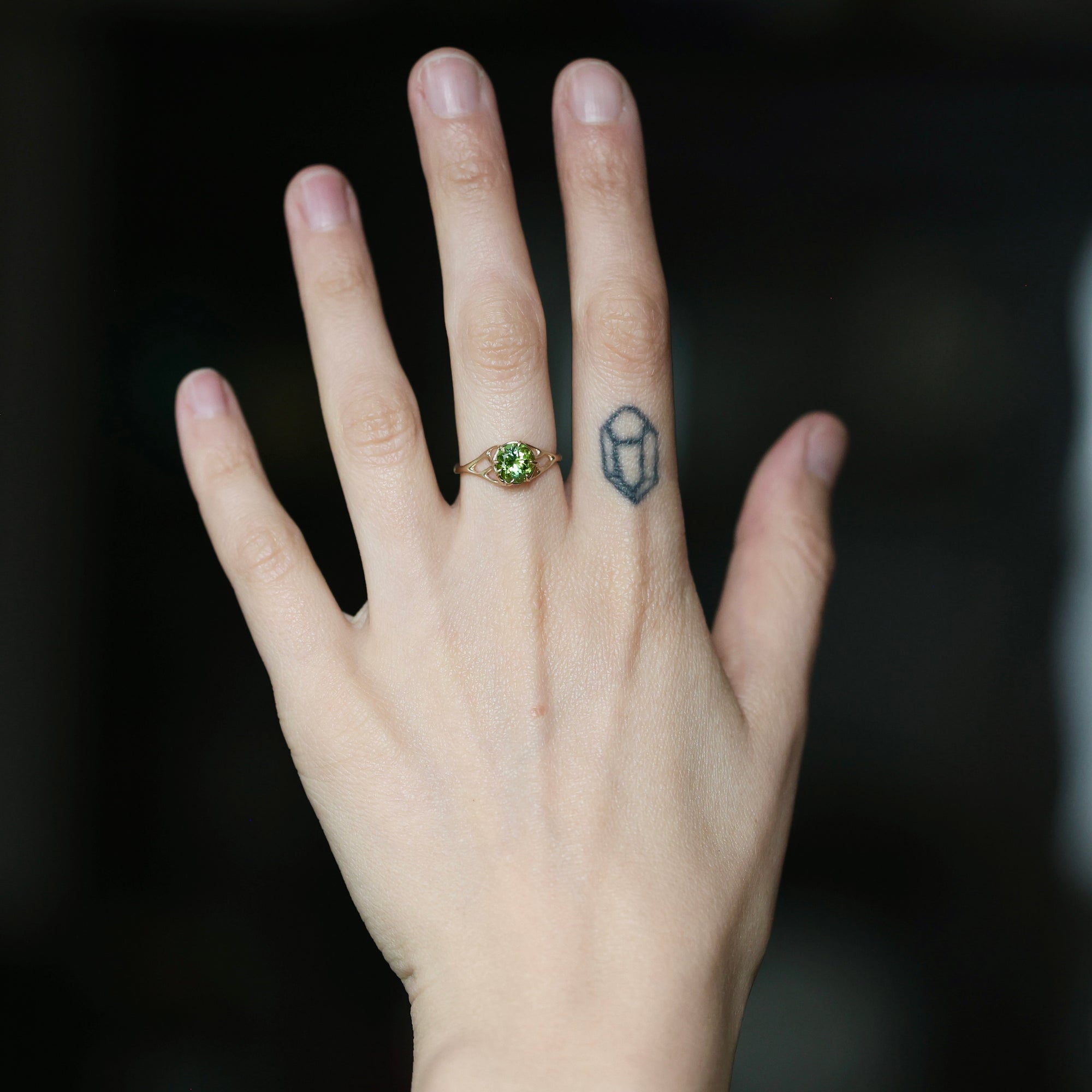 A close-up of a person's hand wearing a gold ring with a green gemstone, set against a dark blurred background.