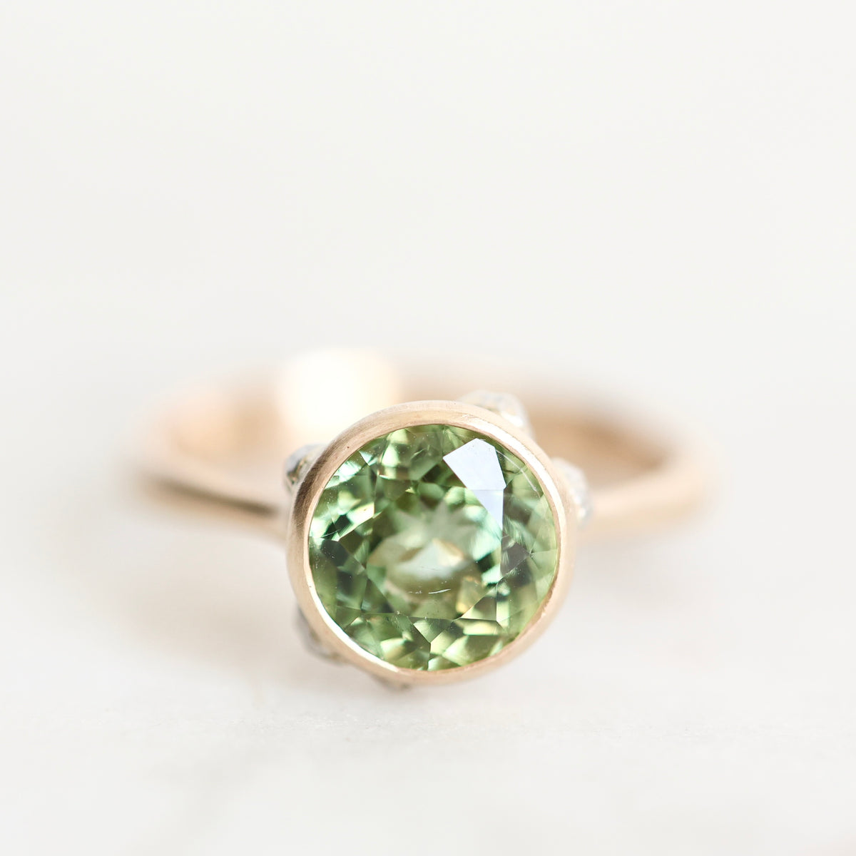 A close-up shot of a ring featuring a round faceted green gemstone in a rose gold bezel setting, with a silver-toned band partially visible on either side, set against a clean white background.