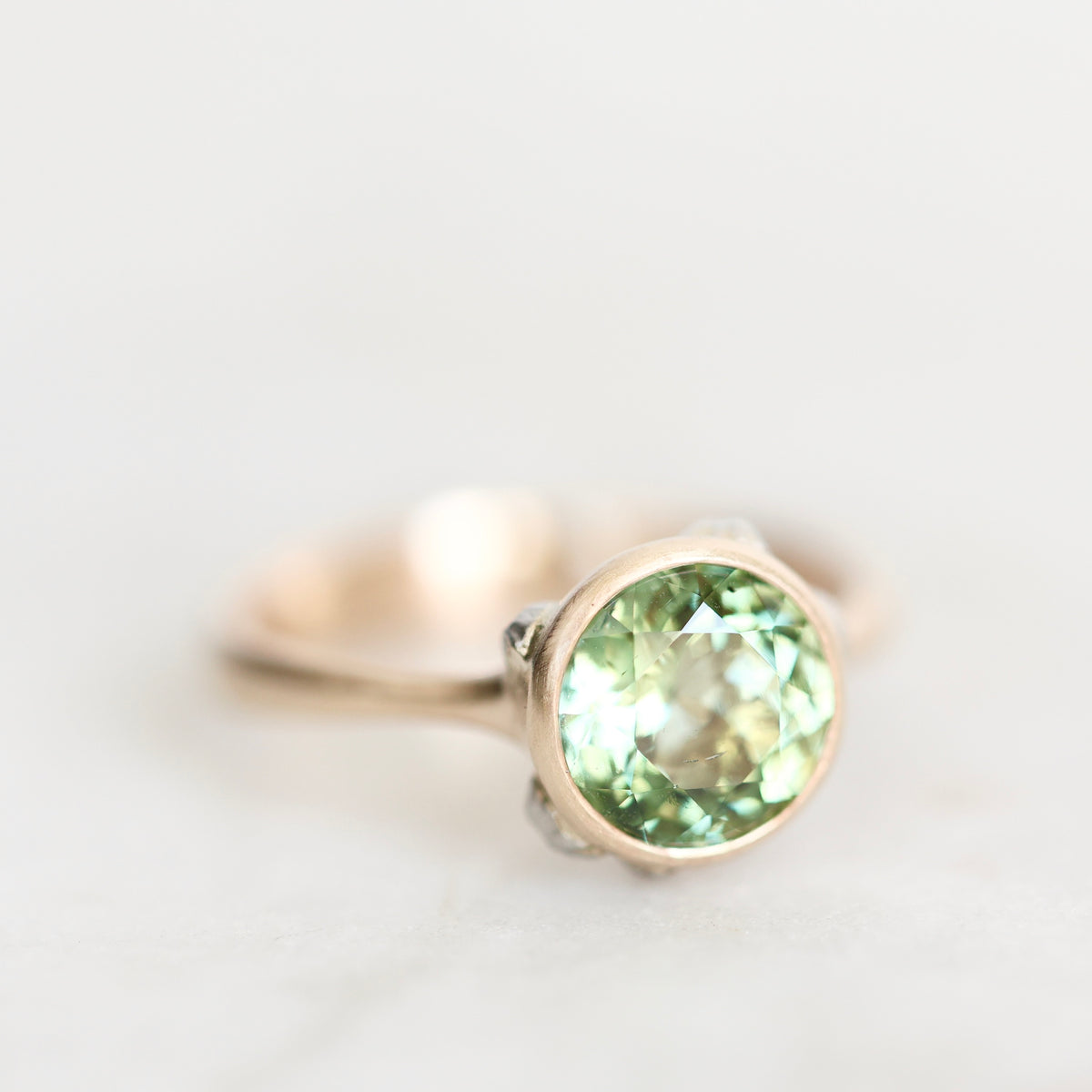 A close-up shot of a ring featuring a round faceted light green gemstone in a rose gold setting, resting on a light, neutral surface.