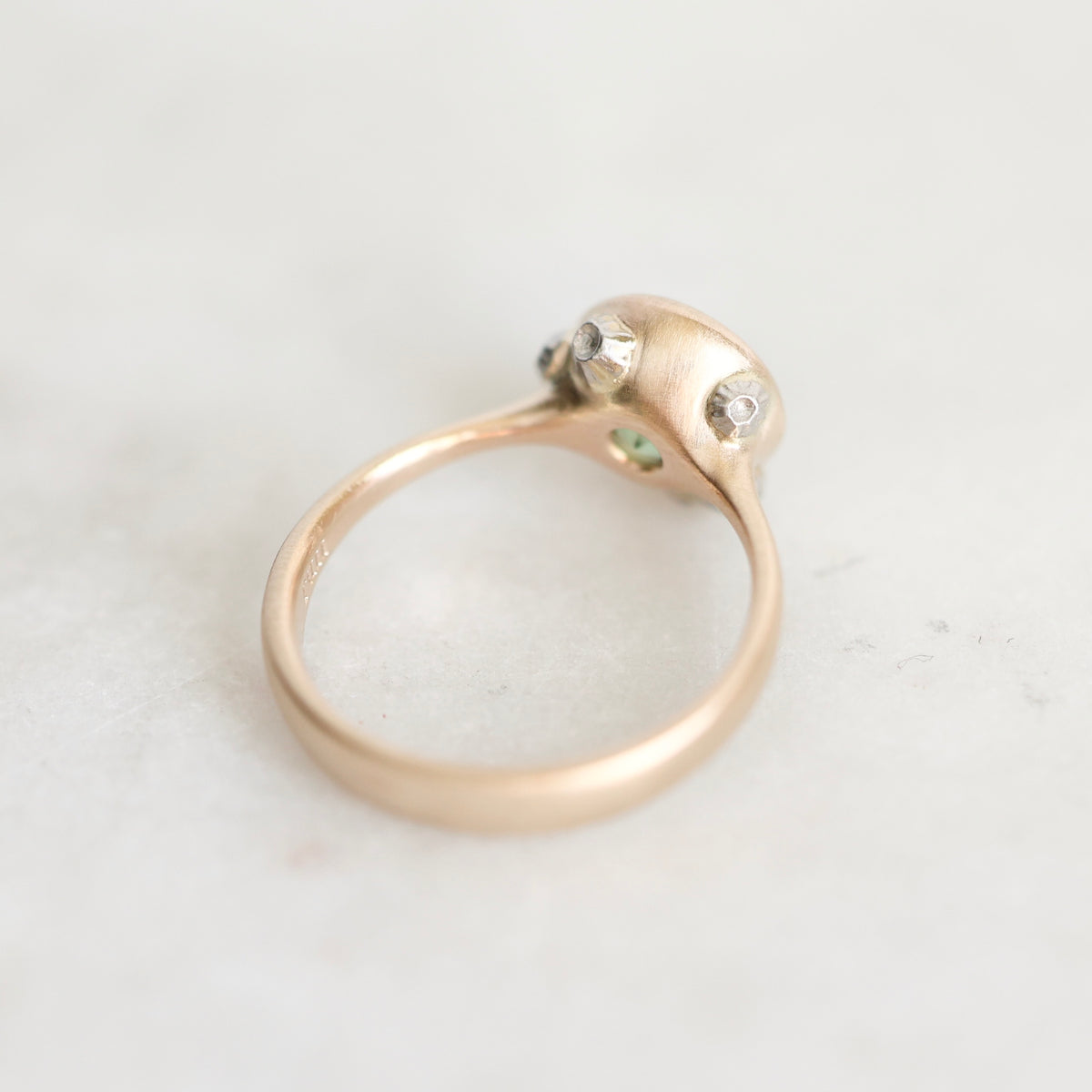 A close-up of a rose gold ring with a textured dome setting featuring two small clear stones and tiny green accents, resting on a light marble surface.