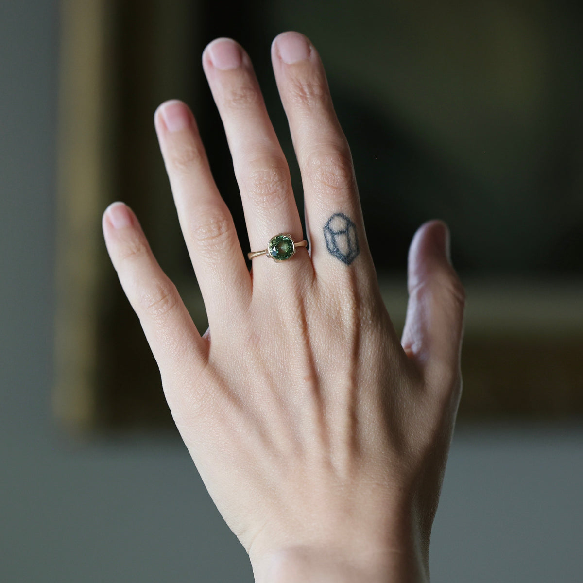 A close-up of a person's hand wearing a gold ring with a round green gemstone, with a small geometric tattoo on the middle finger, against a softly blurred dark background.