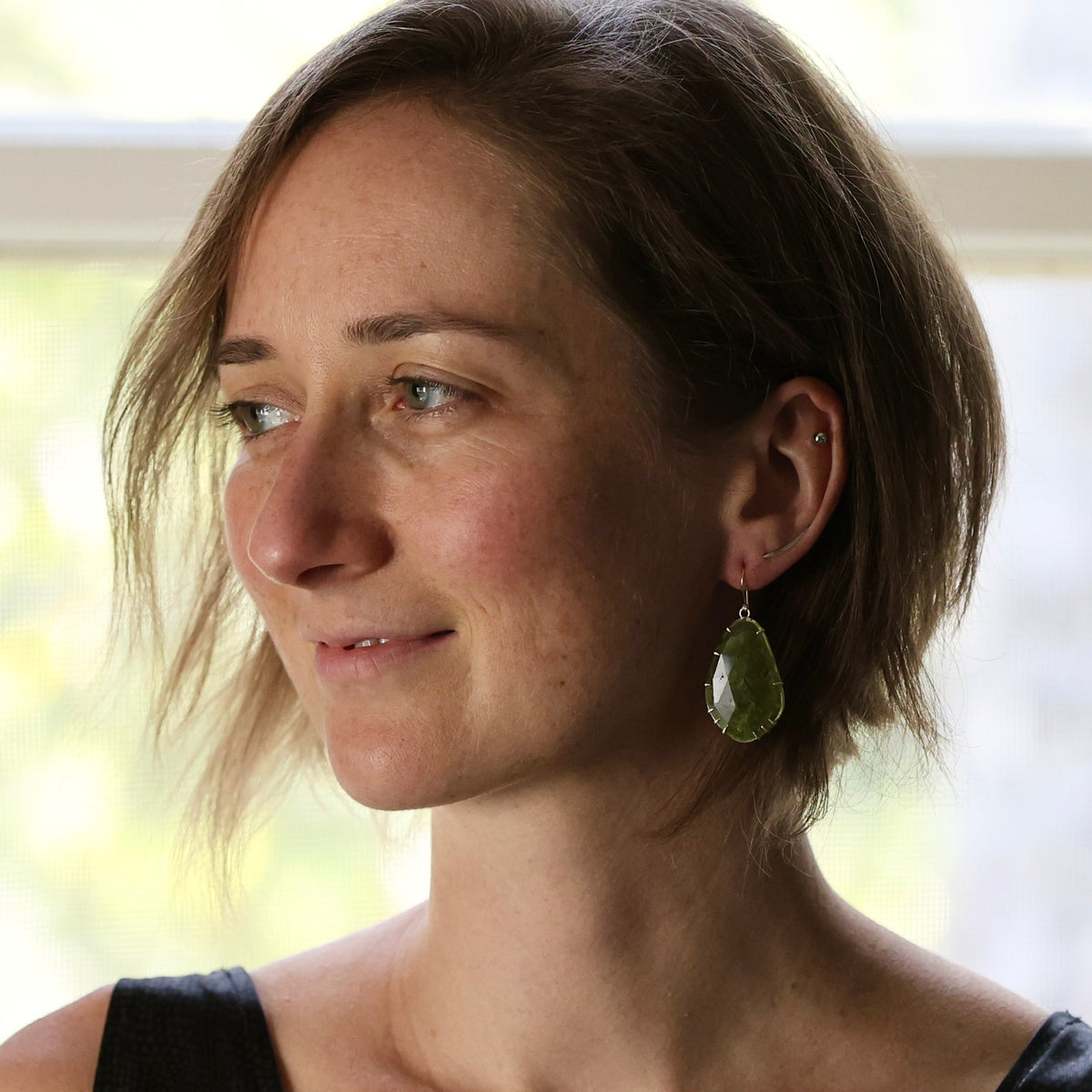 A woman with short brown hair is shown in profile, wearing a dangling green teardrop-shaped earring. The photo is taken indoors near a bright window, with soft natural light and a blurred light background.
