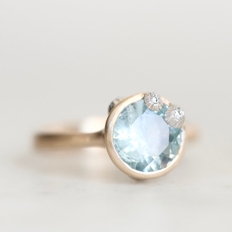 A close-up shot of a ring featuring a large round light blue gemstone in a gold bezel setting, with two small clear accent stones on the upper right, set against a soft, light gray background.