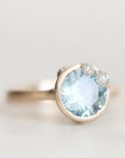 A close-up shot of a ring featuring a large round light blue gemstone in a gold bezel setting, with two small clear accent stones on the upper right, set against a soft, light gray background.