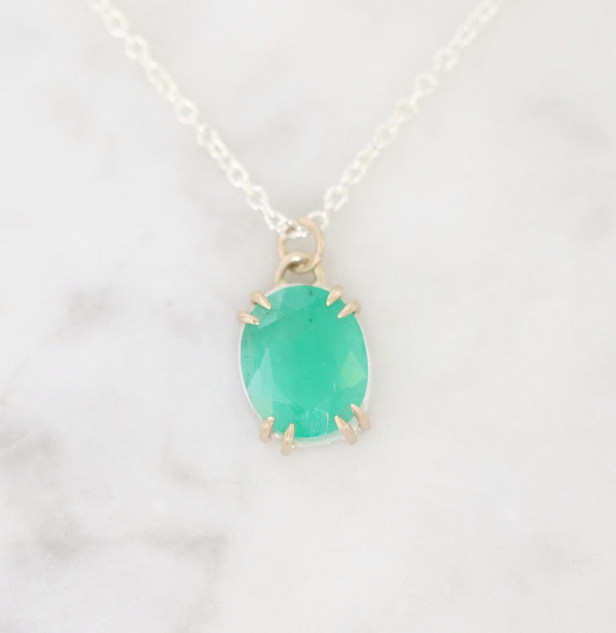 An oval-cut green gemstone pendant in a gold prong setting on a delicate silver chain, displayed on a white marble surface.