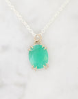 An oval-cut green gemstone pendant in a gold prong setting on a delicate silver chain, displayed on a white marble surface.