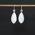 A pair of teardrop-shaped white moonstone drop earrings with gold hooks, hanging from a gold display bar against a solid black background.