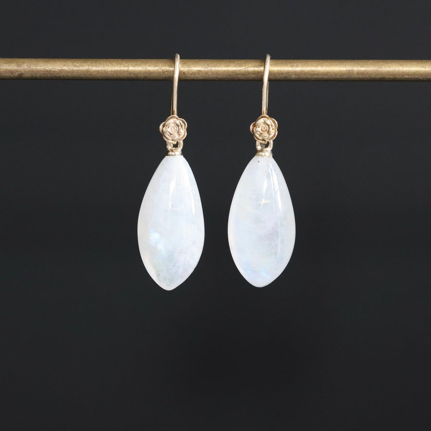 A pair of teardrop-shaped white moonstone drop earrings with gold hooks, hanging from a gold display bar against a solid black background.