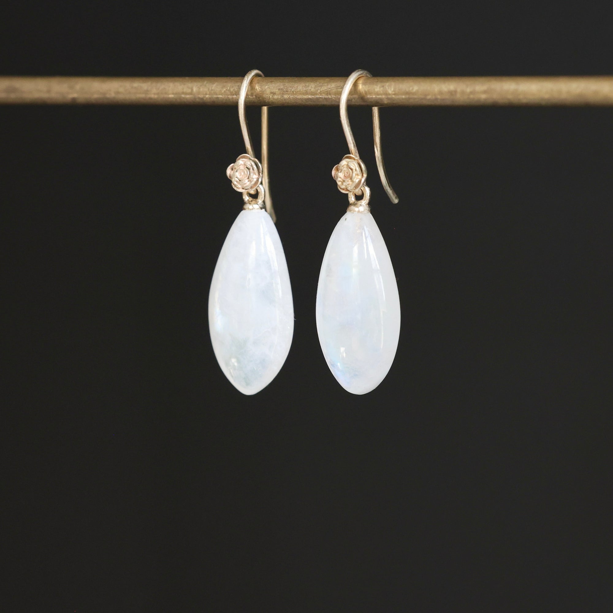 A pair of teardrop-shaped white iridescent gemstone drop earrings with gold hooks, hanging from a gold display bar against a solid black background.