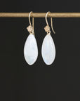 A pair of teardrop-shaped white iridescent gemstone drop earrings with gold hooks, hanging from a gold display bar against a solid black background.